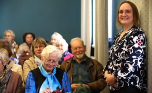 Seniors sit in the large hall of the Südalinna library waiting for the presentation of Jaak Aru's book Alla 65. keelatud to begin. In the foreground sits an old woman fumbling with her phone and an older man smiling at the camera just behind her. Leelo, the cheerful librarian, stands against the wall on the right.